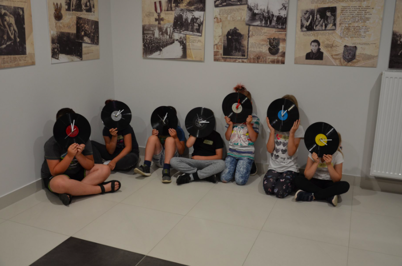 Grupa dzieci siedzących na podłodze. W dłoniach trzymają płyty gramofonowe, zasłaniają nimi twarze.