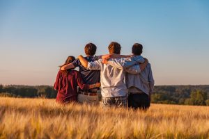 Grupa młodych osób stoi w zbożu tyłem do fotografa. Trzymają się w uścisku.
