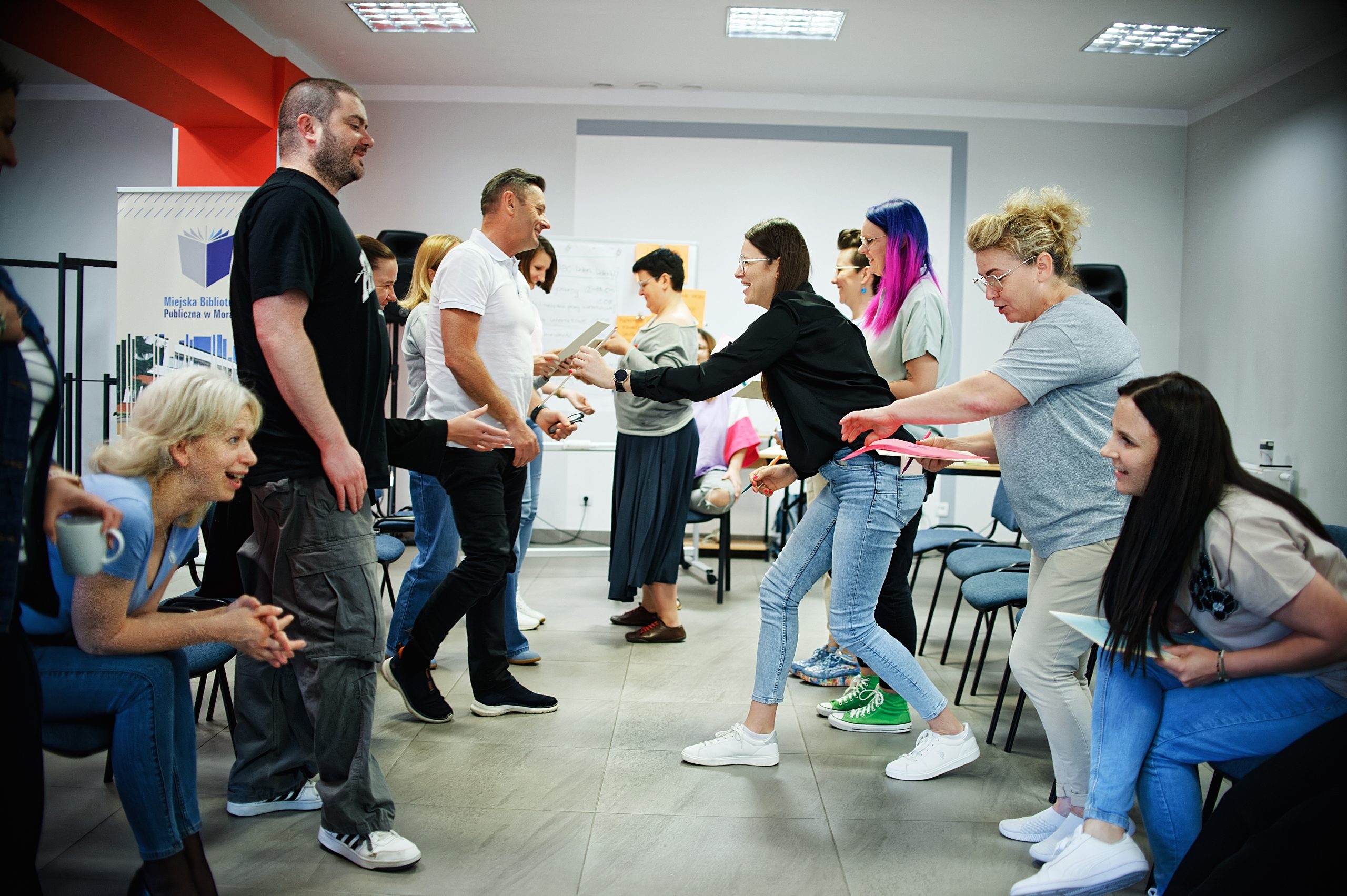 Grupa osób stojących i siedzących naprzeciwko siebie. Część wręcza sobie kartki z portretami.