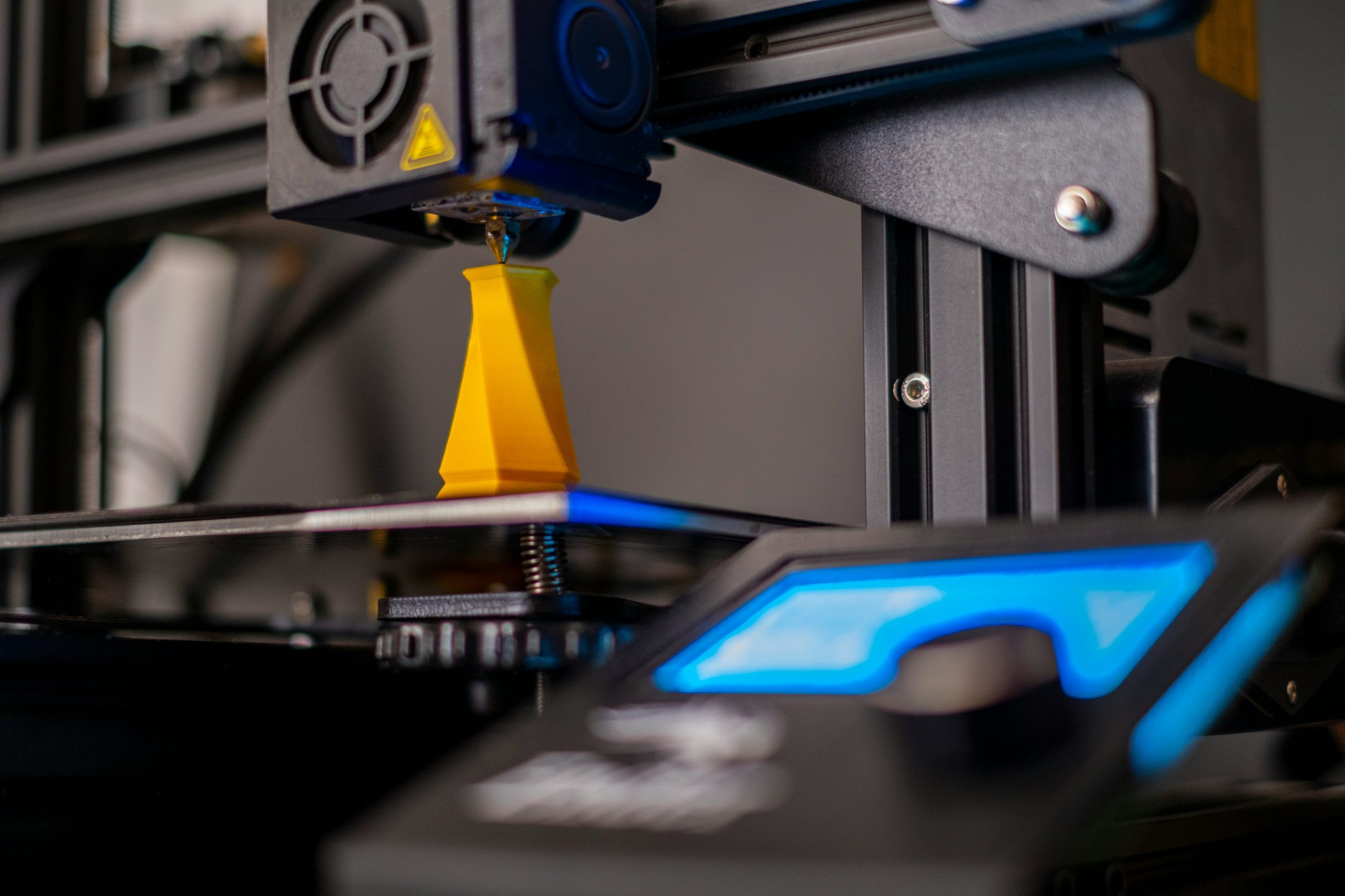 Drukarka 3D drukująca element z żółtego filamentu.
