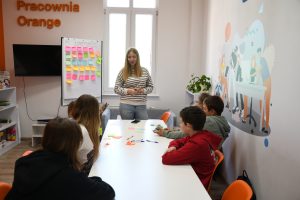 Grupa dzieci siedzi przy stole i słucha stojącej przed nimi kobiety.