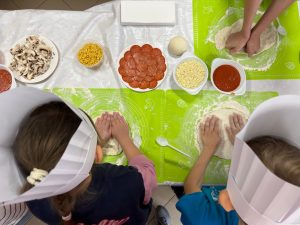 widok z lotu ptaka na stół na którym dzieci wyrabiają ciasto na pizzę. na stole widać sosy i dodatki do pizzy.