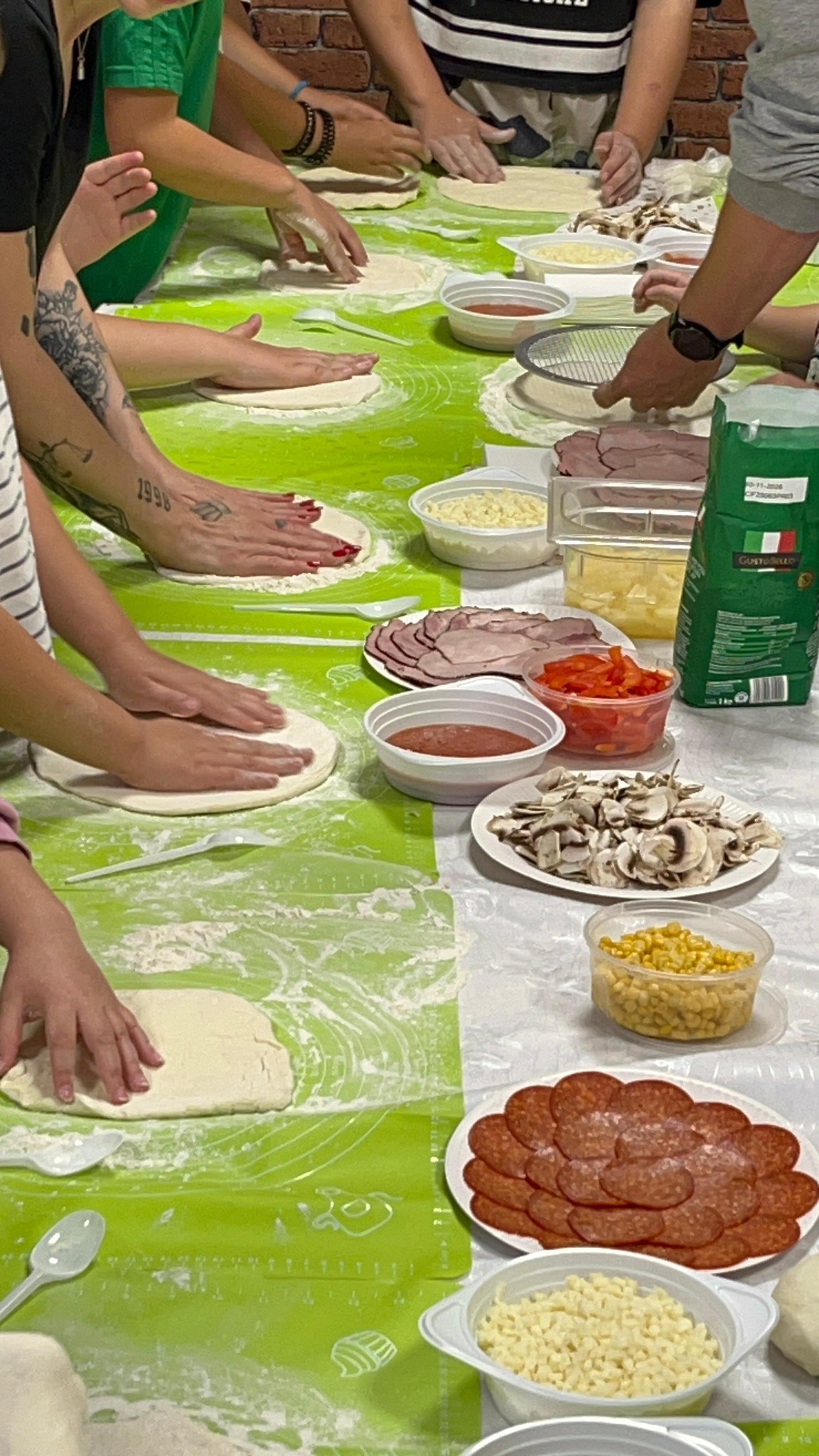 widok na stół na zielony stół na którym dzieci wyrabiają ciasto na pizzę. na stole widać sosy i dodatki do pizzy.