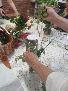Stół zastawiony roślinami i kwiatami; podczas warsztatów botanicznych ludzie układają świeże zioła w słoikach.