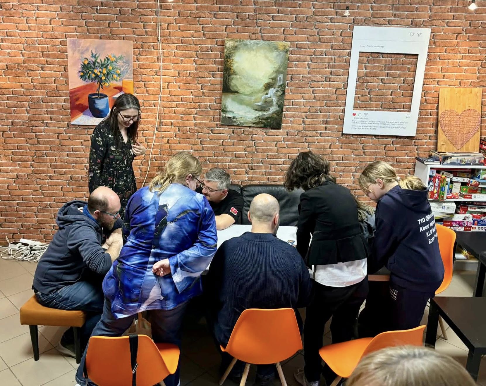 Grupa osób siedzi i stoi wokół stołu podczas spotkania w CAL Pracownia Orange. W tle ceglana ściana, obrazy, regał z grami planszowymi i pomarańczowe krzesła.