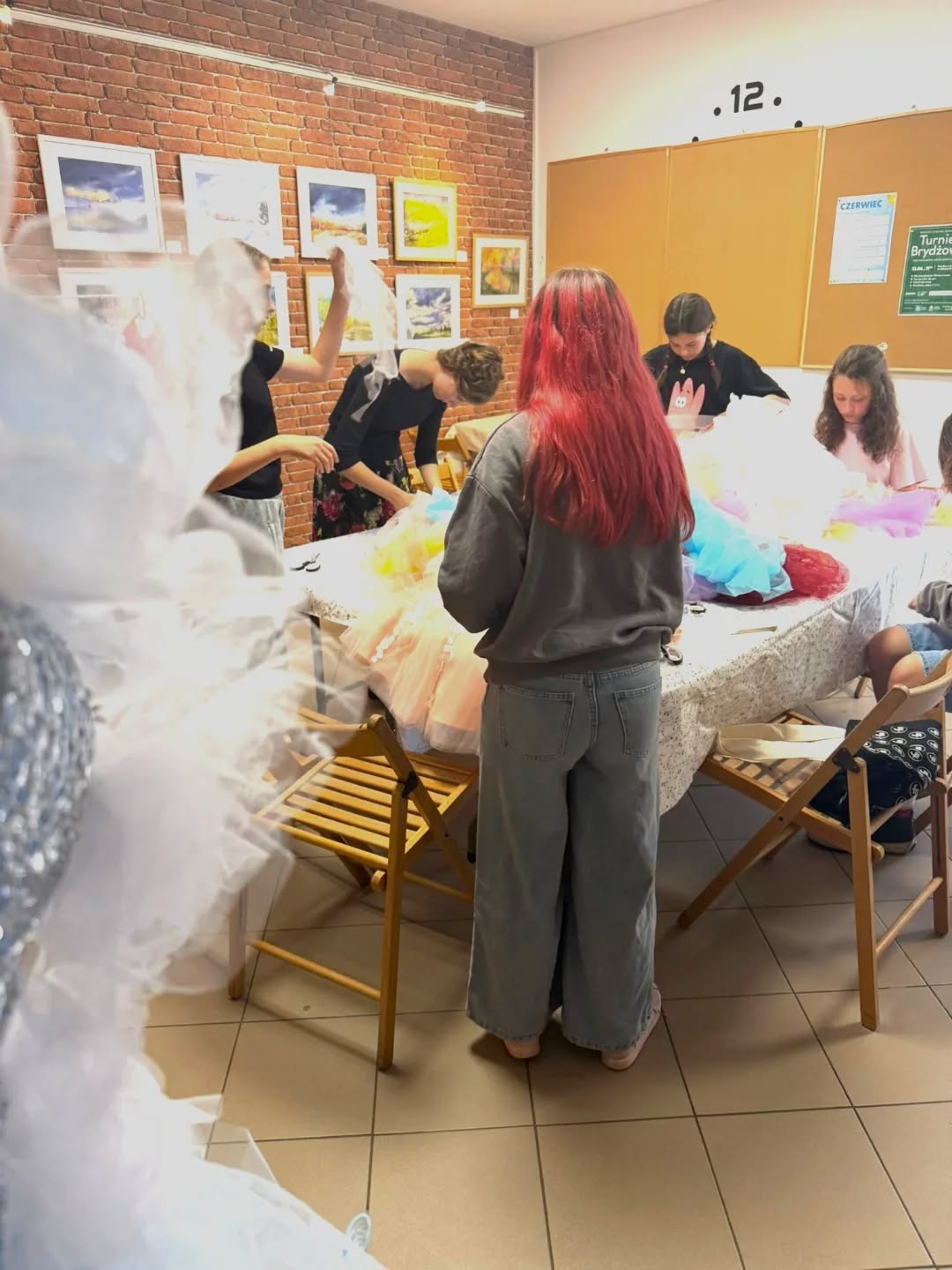 Kilka osób stoi i siedzi wokół dużego stołu w sali z ceglaną ścianą. Na stole leżą kolorowe kawałki tiulu i materiały krawieckie. Uczestniczki przygotowują elementy strojów.