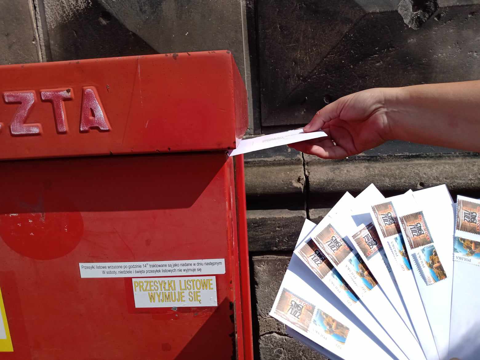 Zdjęcie zaadresowanych kopert oraz ręki liderki Pracowni Orange wkładającej jedną kopertę do skrzynki pocztowej