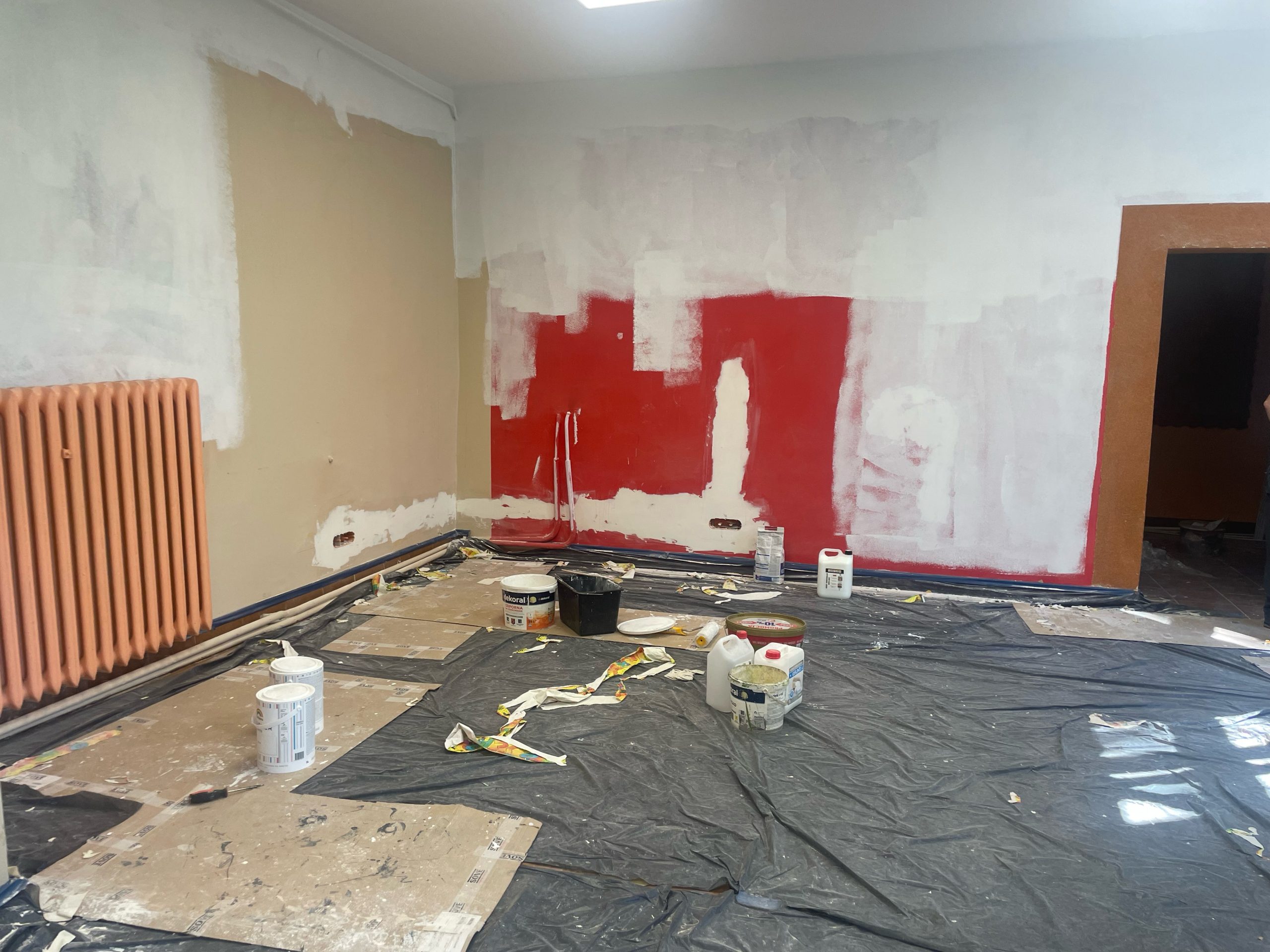 Pomieszczenie w trakcie malowania, czerwona ściana malowana na kolor biały. Na podłodze czarna folia zabezpieczająca i wiadra z farbami.
