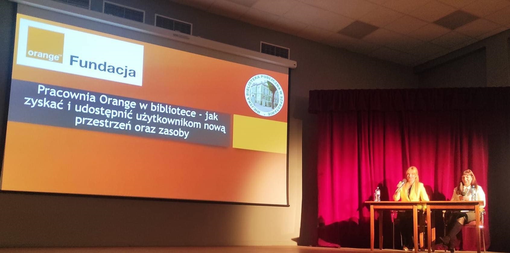 zdjęcie liderek na scenie oraz widoczny slajd z napisem pracownia orange w bibliotece - jak zyskać i udostępnić użytkownikom nową przestrzeń oraz zasoby, na górze logo pracowni orange i biblioteki w szprotawie