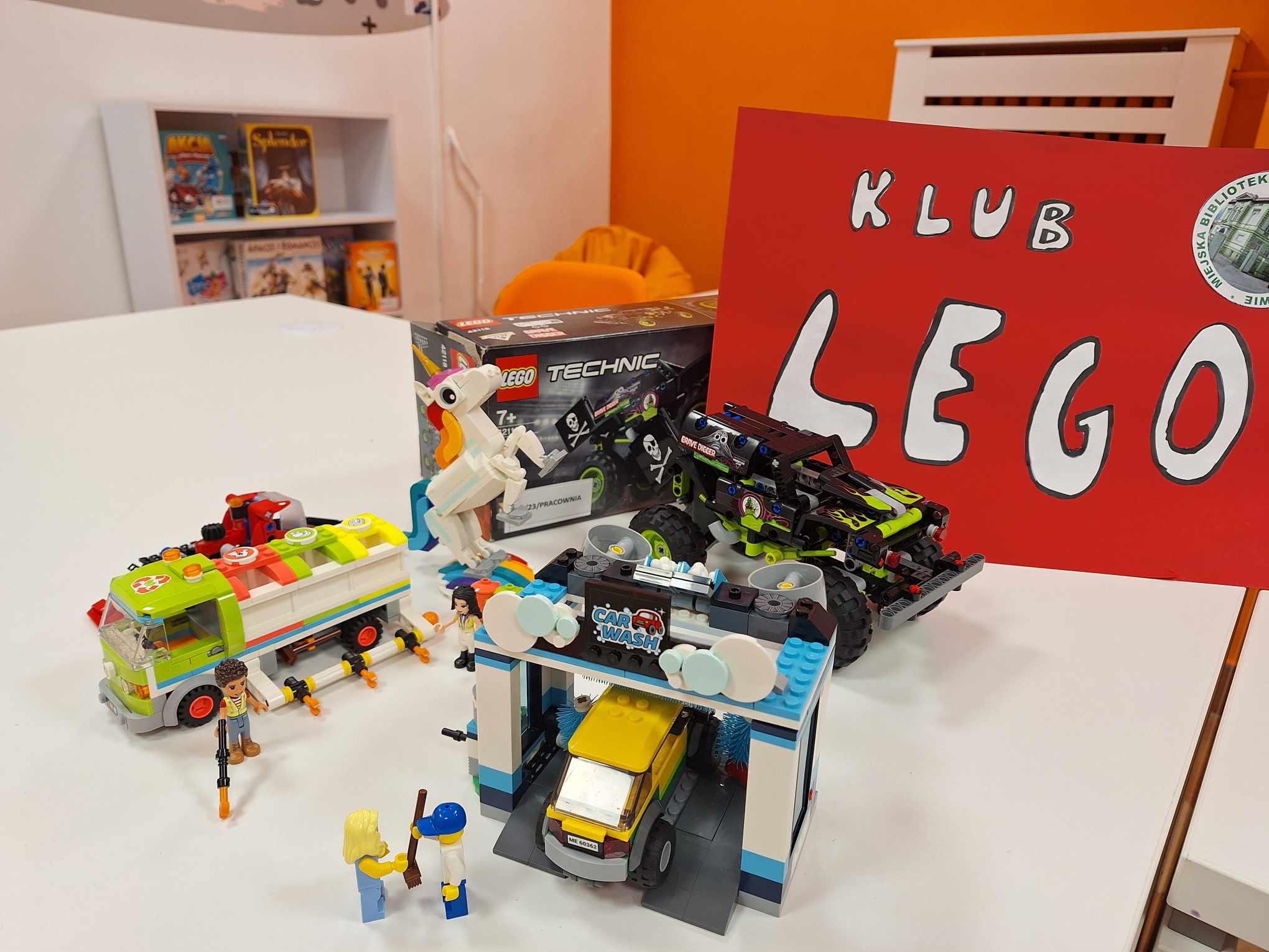 zdjęcie ułożonych modeli myjni samochodowej, śmieciarki i auta terenowego z klocków lego, z tyłu plakat klub lego i logo biblioteki