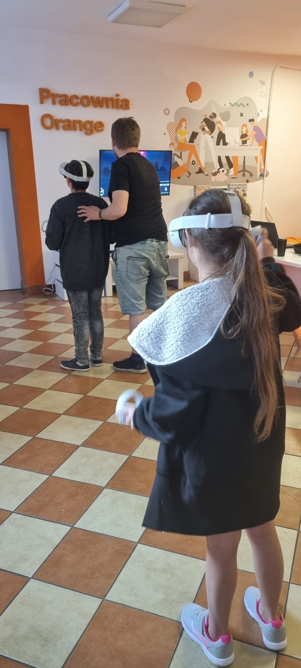 zdjęcie przedstawiające dziewczynkę z goglami VR podczas malowania wirtualnego bałwanka, w tle chłopiec z goglami VR i informatyk tłumaczący działanie aplikacji