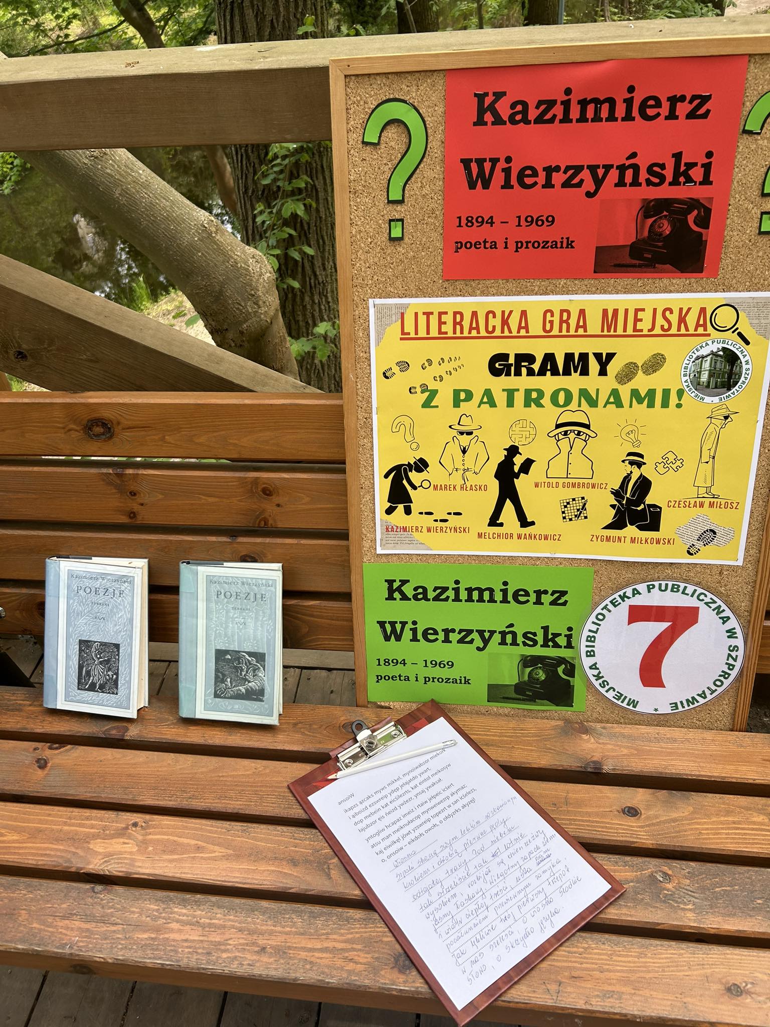 zdjęcie tablicy korkowej wraz z danymi autora Kazimierza Wierzyńskiego oraz plakatem literackiej gry miejskiej, obok książki i podkładki z zadaniami na ławce