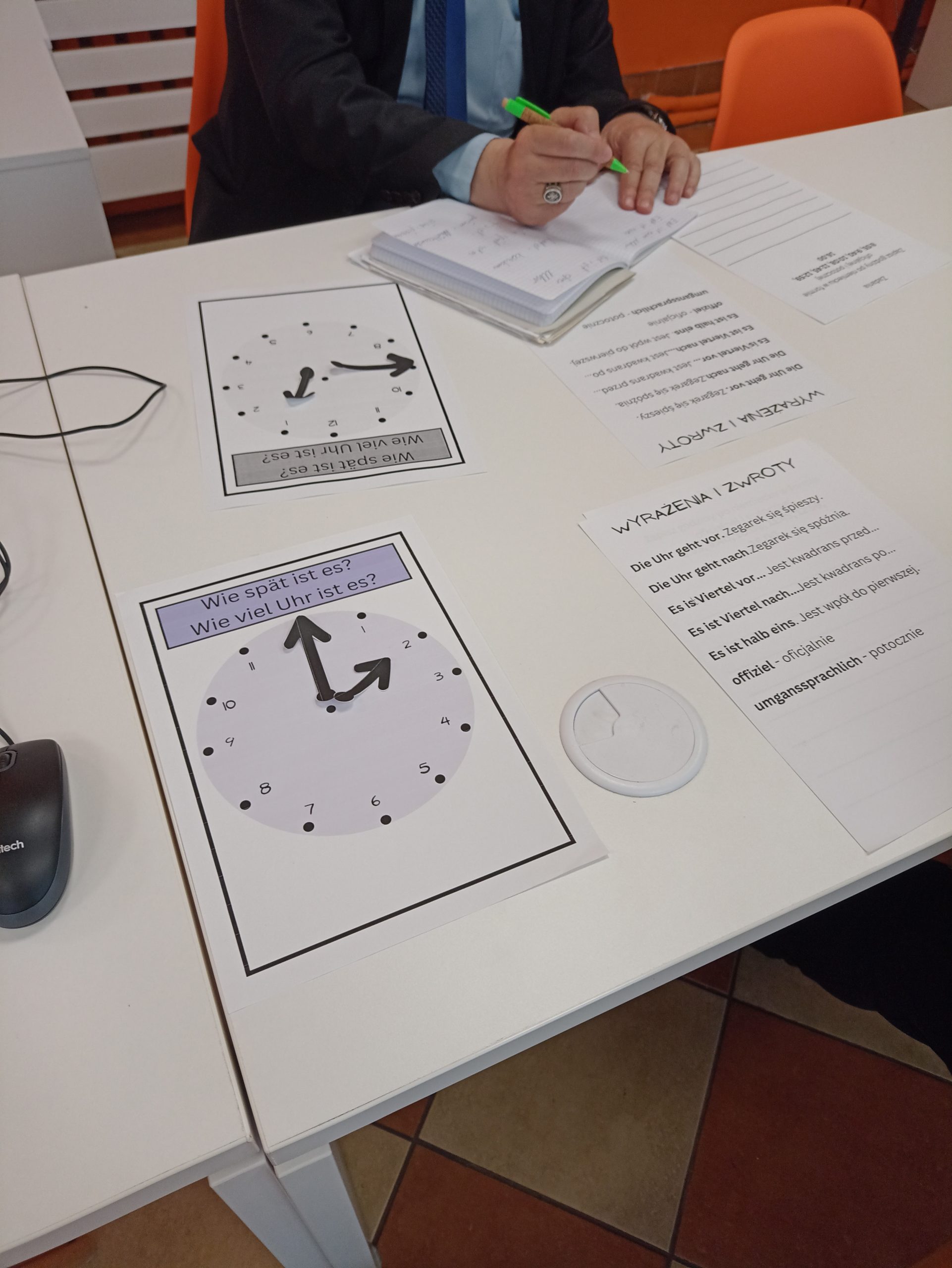 Na stole leżą karty pracy, na jednej grafika przedstawiająca analogową tarczę zegara bez wskazówek, na drugiej są wyrażenia i zwroty po niemiecku.