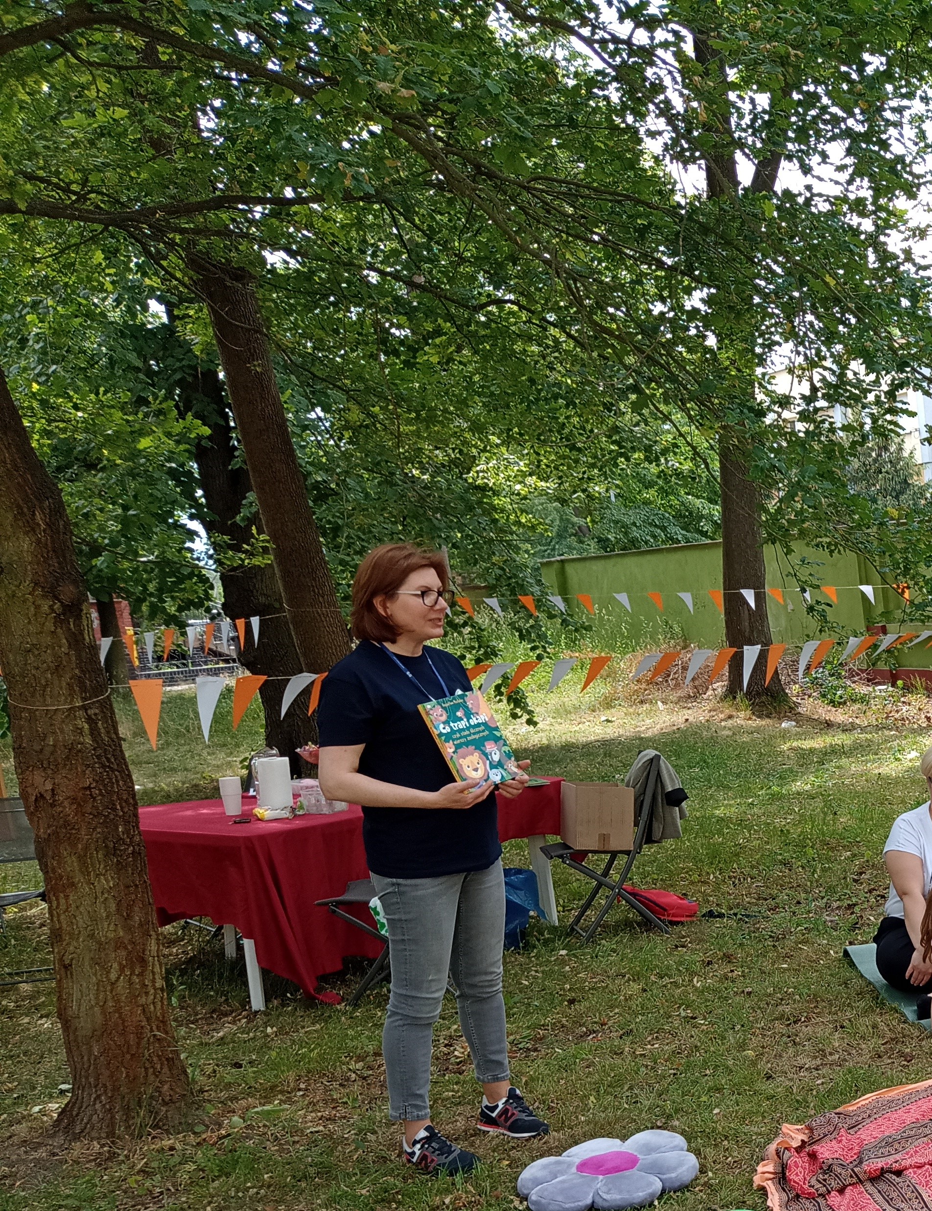 Bibliotekarka stoi na dworze, trzyma w rękach książkę, za nią stół i krzesła, do drzew przyczepiona girlanda z białych i pomarańczowych trójkątów.