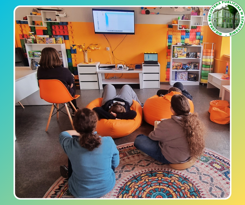 zdjęcie klubowiczów i bibliotekarki podczas oglądania filmu animowanego na telewizorze, po prawej logo biblioteki