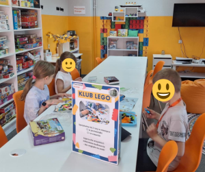 zdjęcie dzieci przy stole podczas składania klocków LEGO, przed nimi plakat o zajęciach