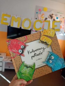 Na zdjęciu widać rękę trzymającą książkę "Kolorowy potwór", w tyle widać żółty napis "emocje"