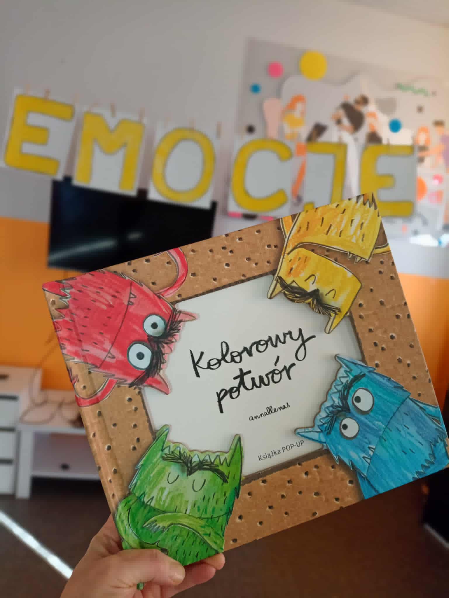 Na zdjęciu widać rękę trzymającą książkę "Kolorowy potwór", w tyle widać żółty napis "emocje"