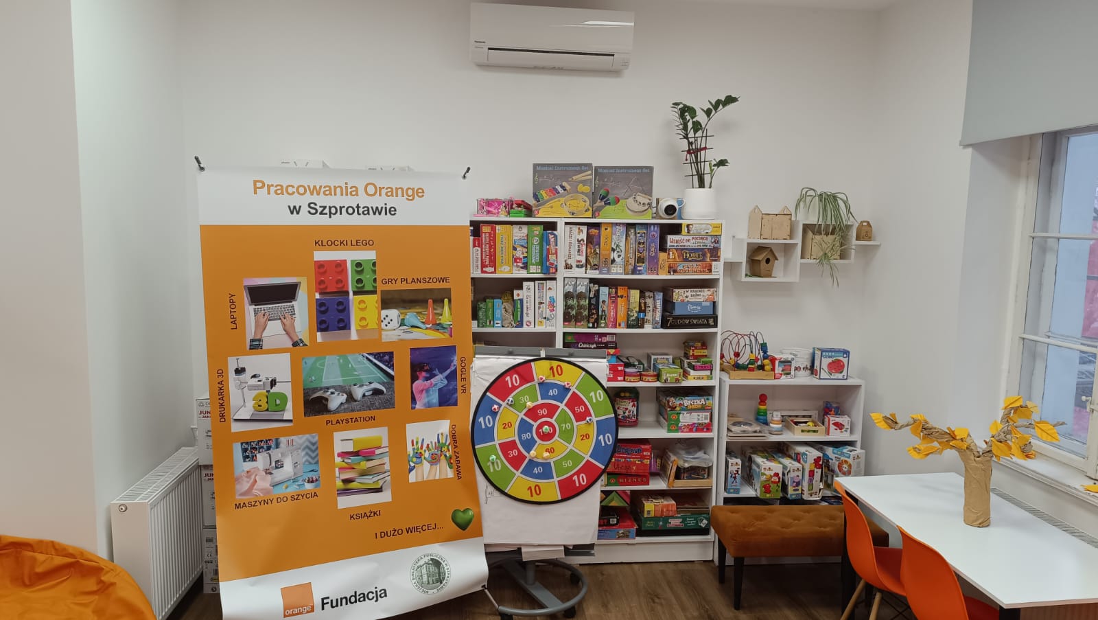 widok na rollup promujący pracownię orange, obok regały wypełnione grami planszowymi , stolik z białym blatem a na nim pomarańczowe papierowe drzewko z pomarańczowymi listkami origami.