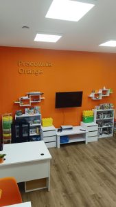 widok na pomarańczową ścianę, na niej telewizor, obok wiszące półki, napis na ścianie Pracownia Orange, komody z grami i klockami lego, widoczny fragment biurka. Meble białe, drewniana podłoga.