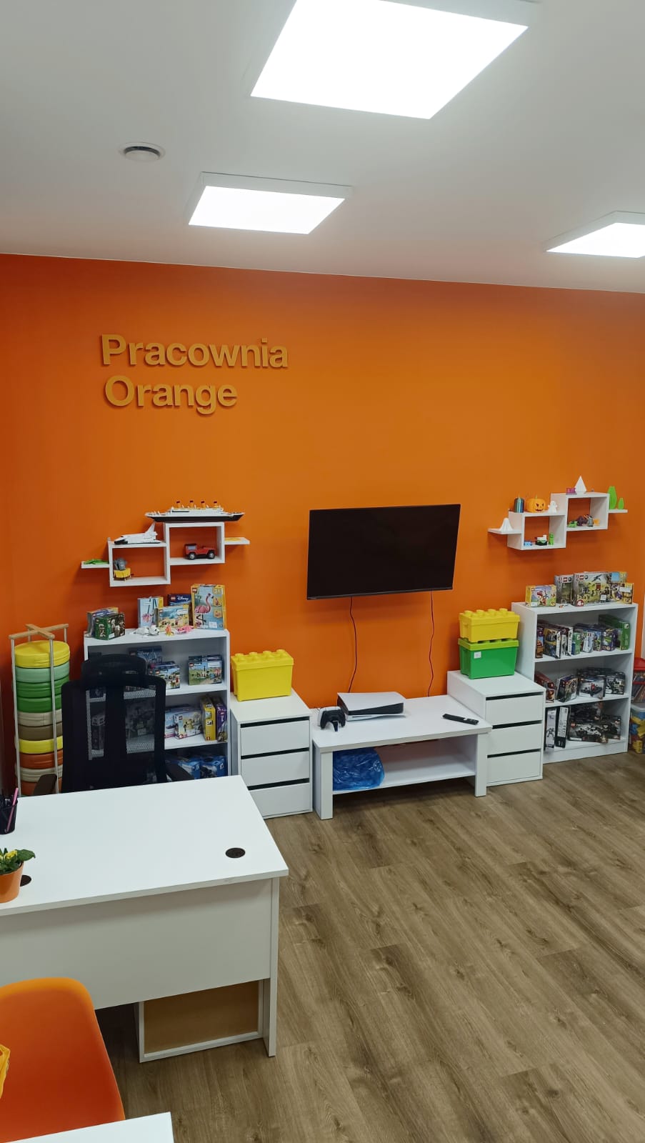 widok na pomarańczową ścianę, na niej telewizor, obok wiszące półki, napis na ścianie Pracownia Orange, komody z grami i klockami lego, widoczny fragment biurka. Meble białe, drewniana podłoga.