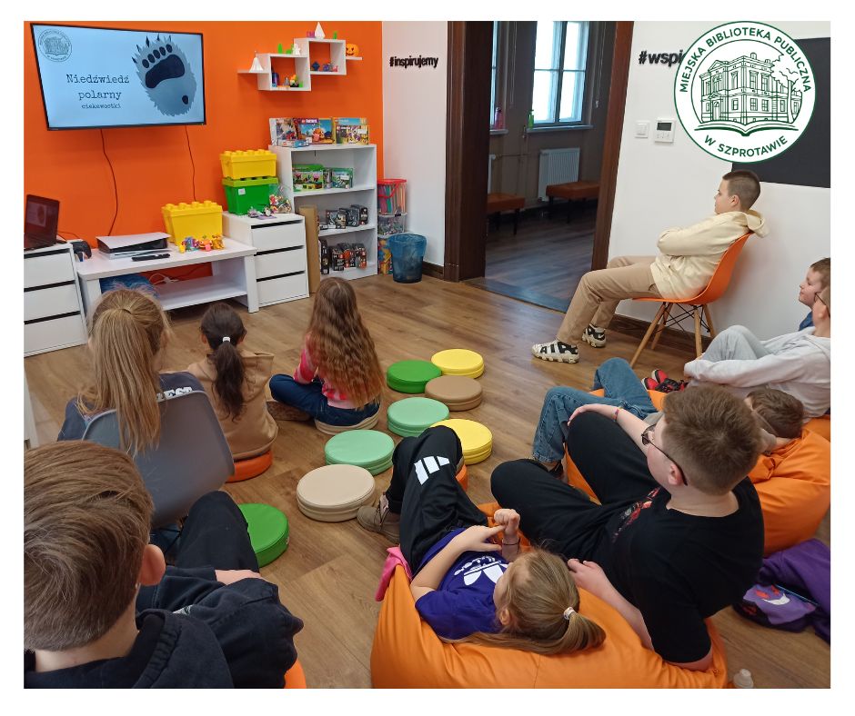 grupa dzieci ogląda prezentację na telewizorze w sali Orange. dzieci siedzą na kolorowych poduszkach i pomarańczowych pufach, na przodzie okrągłe logo Miejskiej Biblioteki Publicznej w Szprotawie.