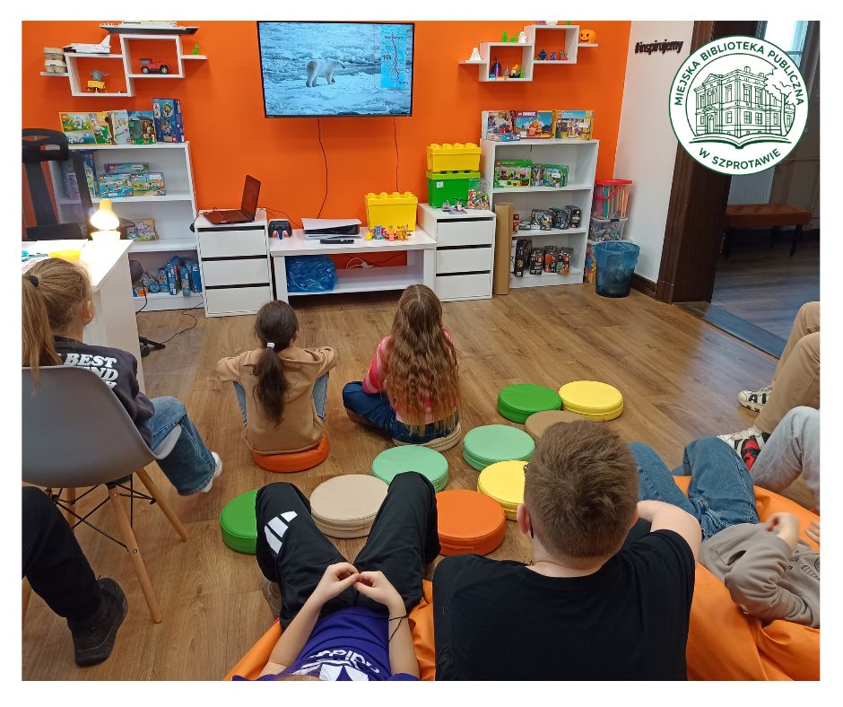 grupa dzieci ogląda prezentację na telewizorze w sali Orange. dzieci siedzą na kolorowych poduszkach i pomarańczowych pufach, na przodzie okrągłe logo Miejskiej Biblioteki Publicznej w Szprotawie.