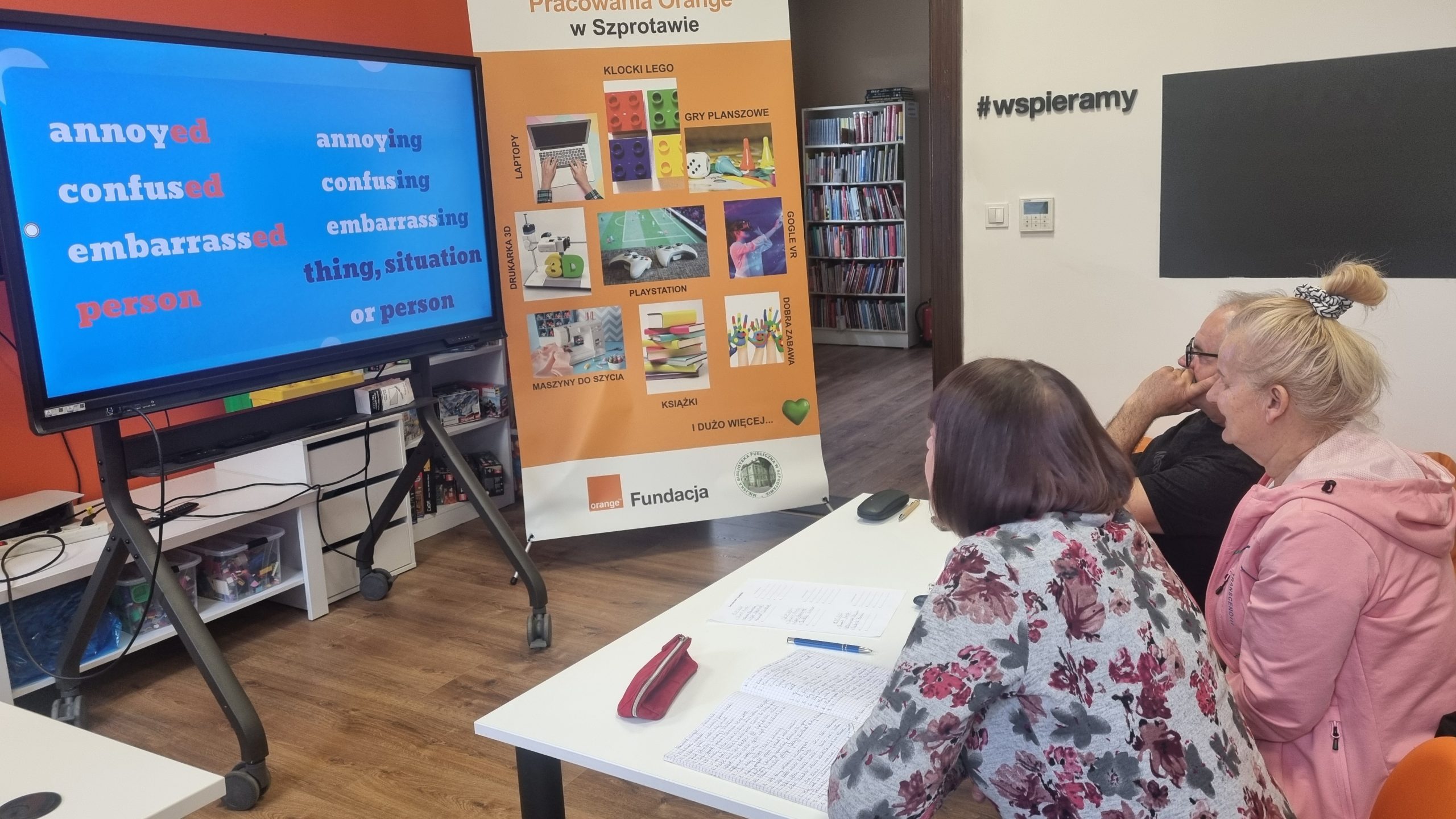 zdjęcie seniorów oglądających filmik edukacyjny na ekranie, w tle baner pracowni orange