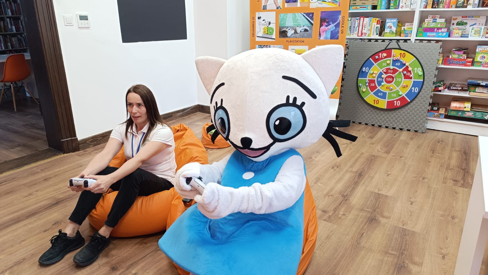 osoba w przebraniu Kici Koci i kobieta siedzą na pomarańczowych pufach i grają w play station, za nimi baner pracowni orange