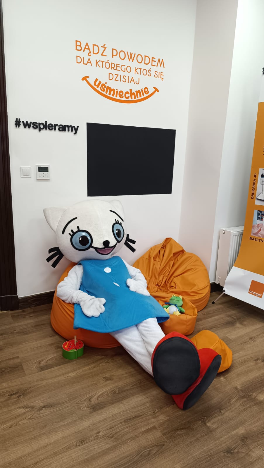 Osoba w przebraniu Kici Koci siedzi na pufie, oparta o ścianę, nad nią cytat na ścianie "Bądź powodem, dla którego ktoś się dzisiaj uśmiechnie".