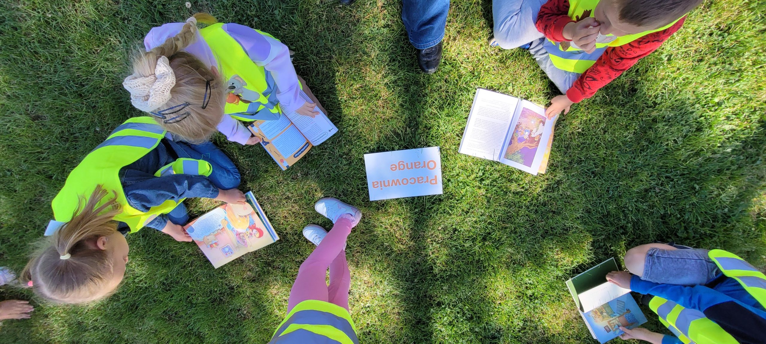 Dzieci siedzą na trawie, czytają książki.