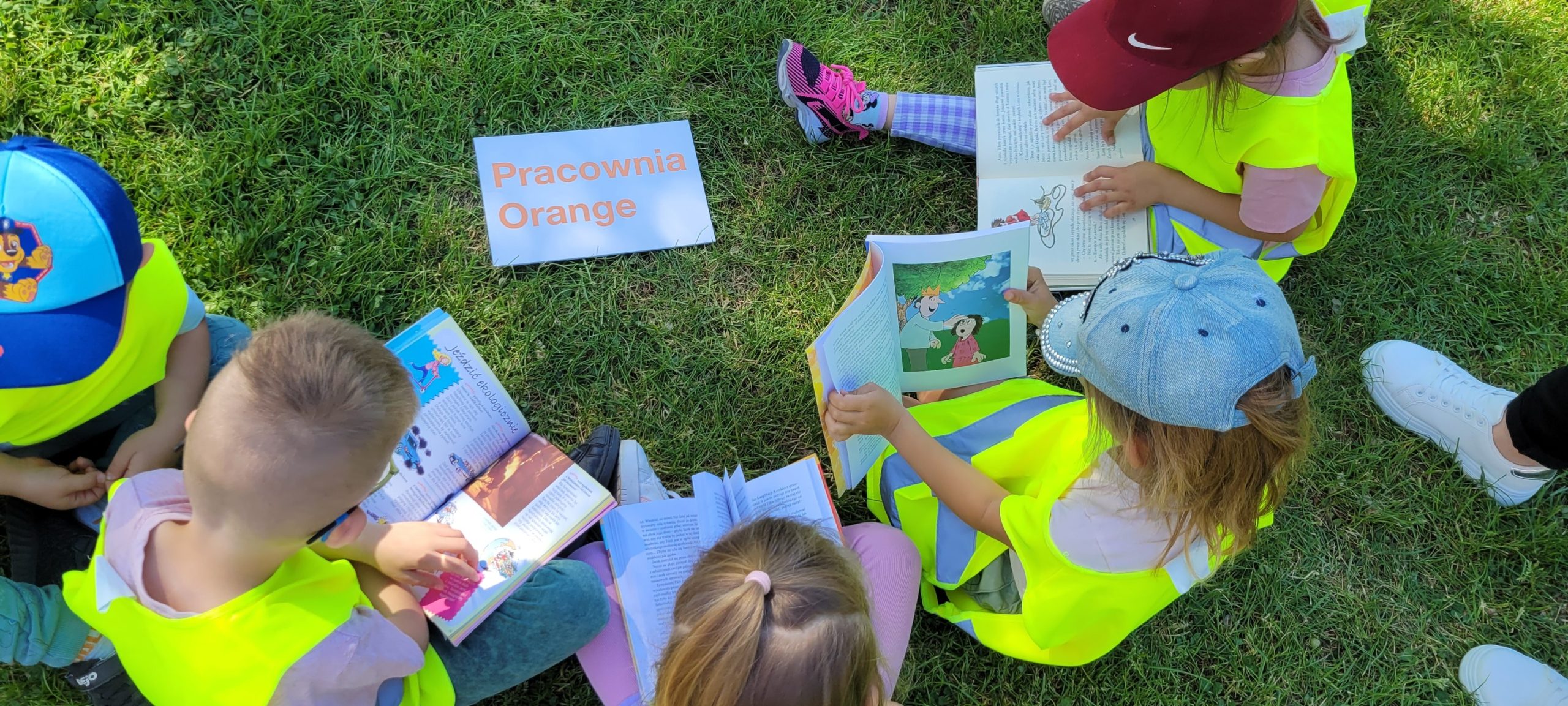 Dzieci siedzą na trawie, czytają książki.