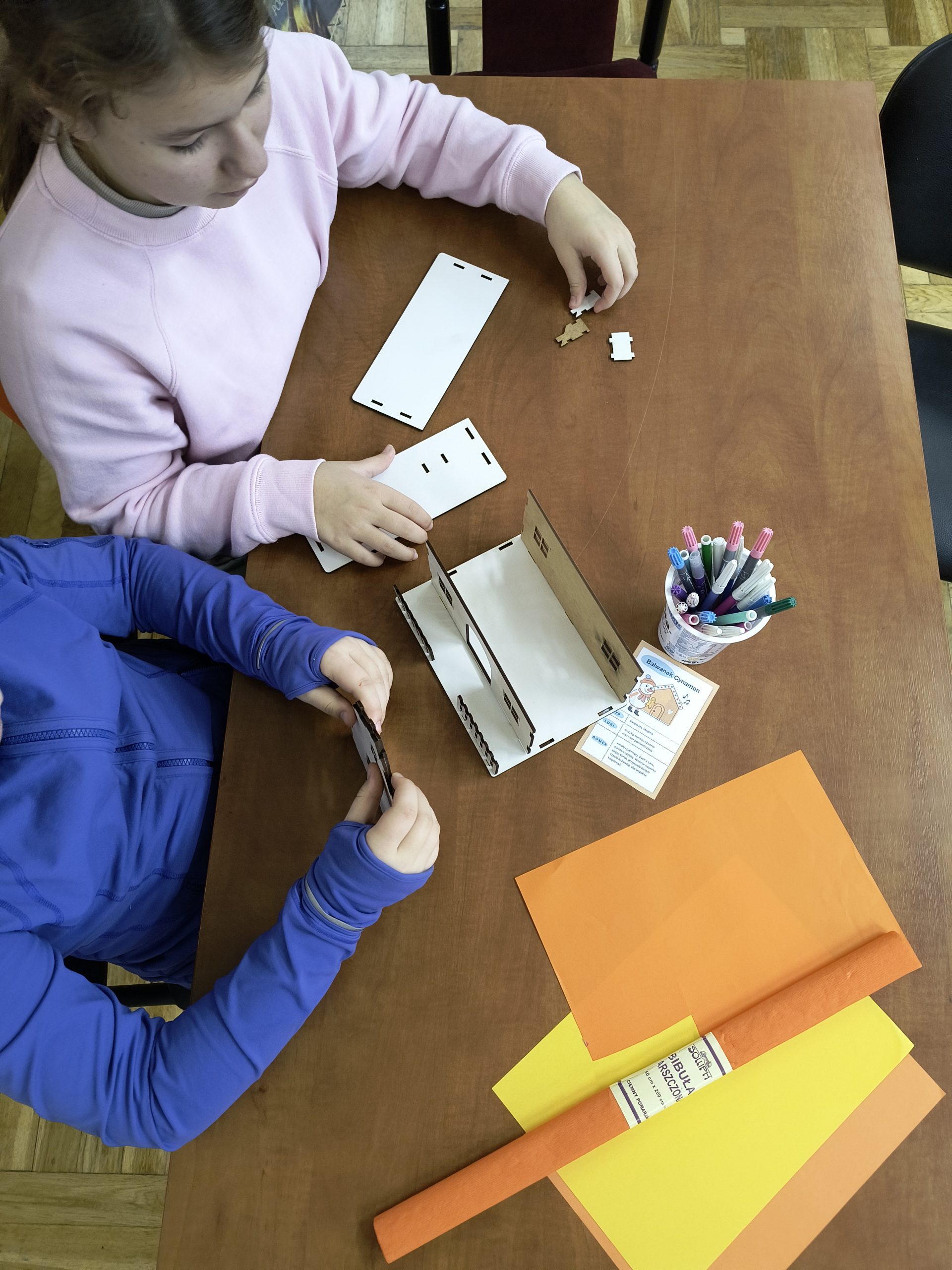 na zdjęciu widać stół z materiałami plastycznymi, dwie osoby siedzące przy stole i składające domek ze sklejki