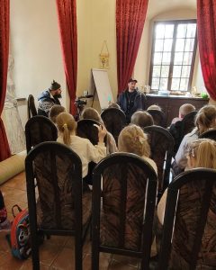 Na zdjęciu widać dzieci siedzące na krzesłach tyłem a przed nimi mężczyznę z kamerą.