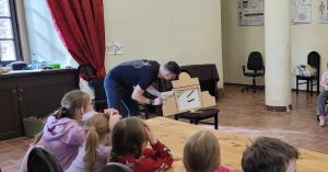 Na zdjęciu widać głowy dzieci z tyłu, przed nimi stół a dalej mężczyznę pochylającego nad krzesłem i pokazującego ramę teatru kamishibai.