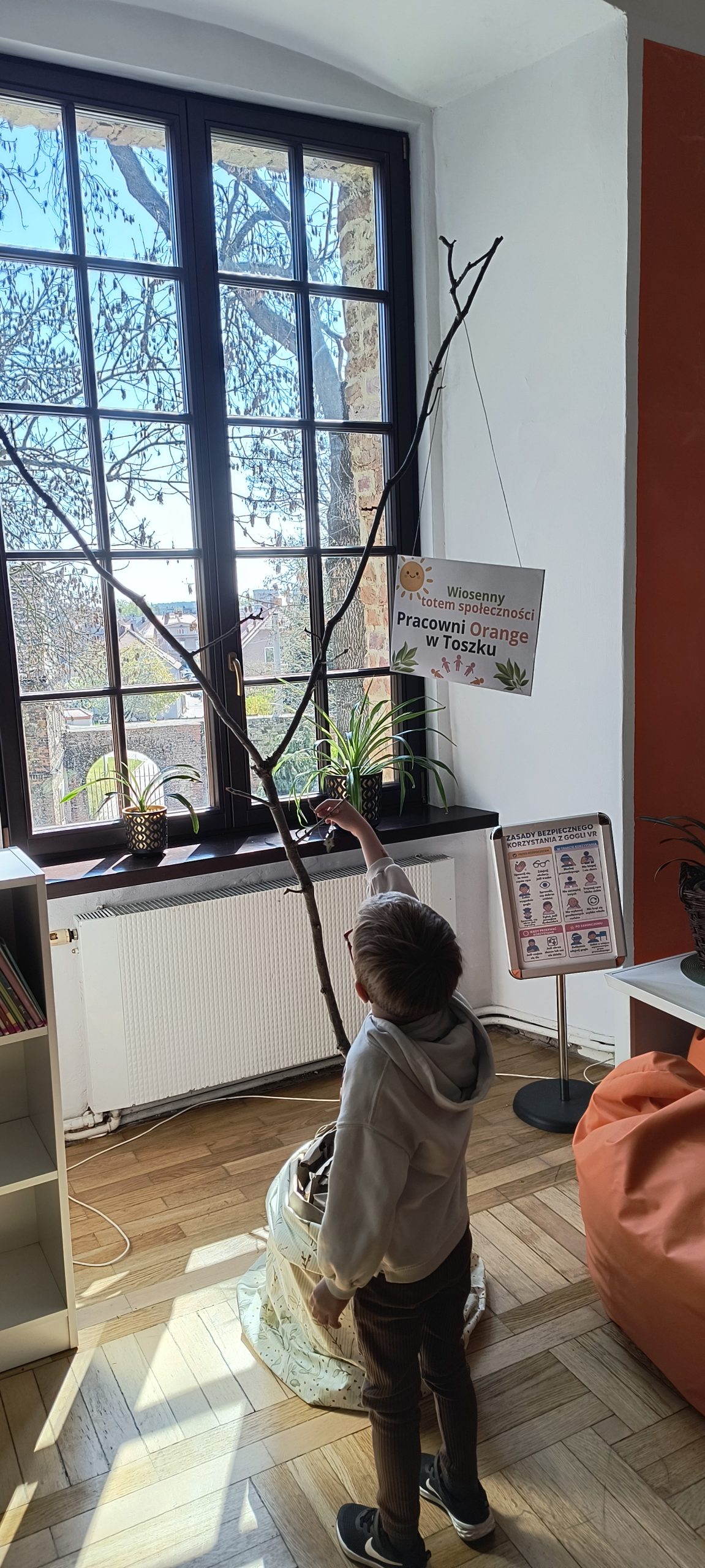Na zdjęciu widać dziecko stojące tyłem przed gałęzią w doniczce, w tle duże okno.