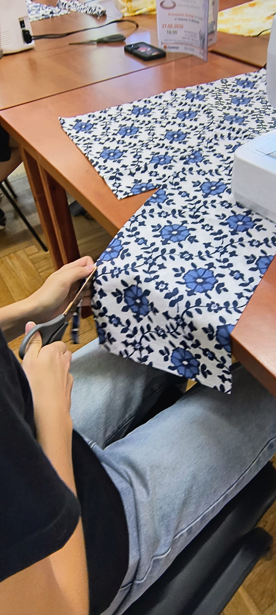 Na zdjęciu widać ręcę oraz nogi osoby siedzącej przy stole, w dłoniach trzyma nożyczki i tenie kawałek kolorowego materiału. Materiał leży na stole.