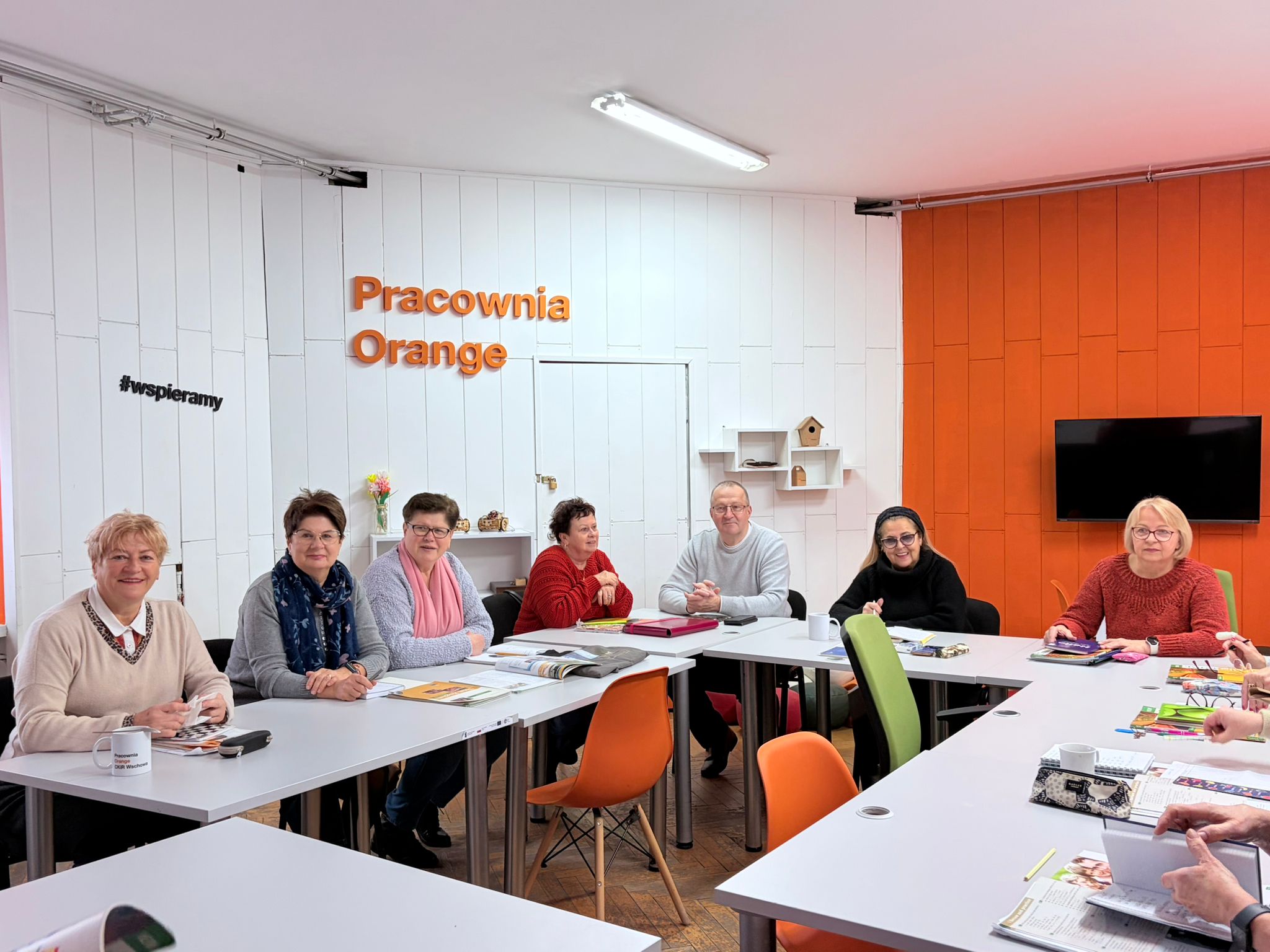 Na zdjęciu znajduje się grupa osób siedzących wspólnie przy dużych, białych stołach. Biorą oni udział w spotkaniu w „Pracowni Orange”. Pomieszczenie jest jasne, uporządkowane i ma charakterystyczne pomarańczowe akcenty. Uczestnicy wyglądają na uśmiechniętych i zaangażowanych w rozmowę.