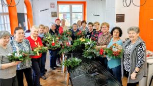 Na zdjęciu znajduje się grupa osób, która prezentuje własnoręcznie wykonane stroiki świąteczne w Pracowni Orange. Widać uśmiechy i radość uczestników podczas świątecznych zajęć.