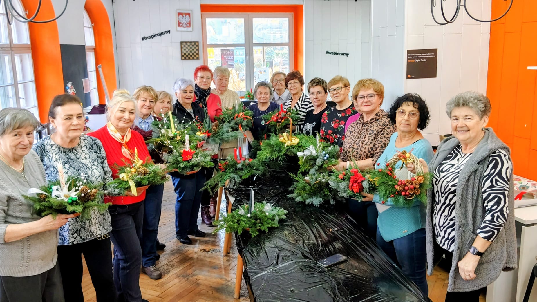 Na zdjęciu znajduje się grupa osób, która prezentuje własnoręcznie wykonane stroiki świąteczne w Pracowni Orange. Widać uśmiechy i radość uczestników podczas świątecznych zajęć.