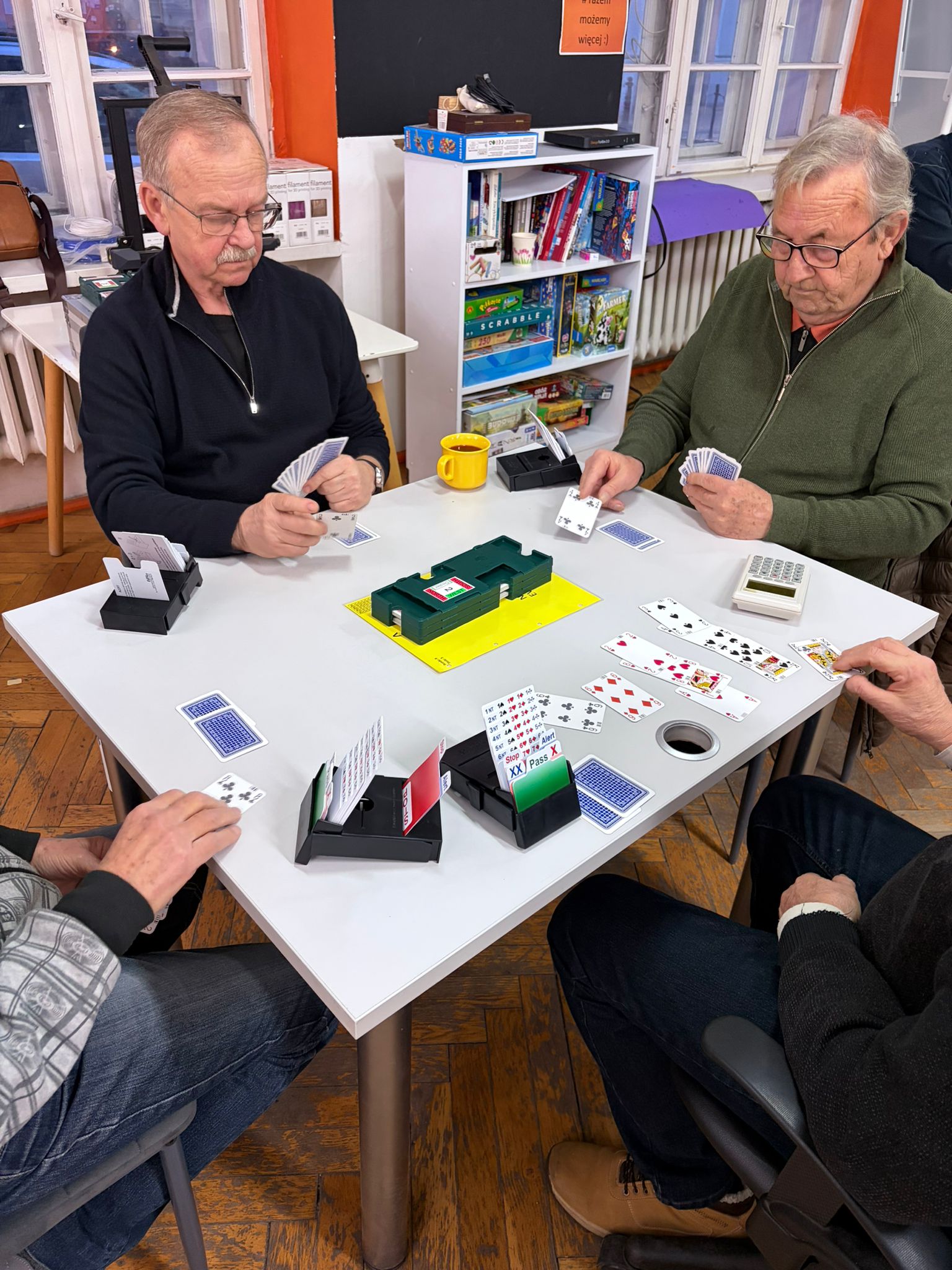 Na zdjęciu znajduję się grupa osób grająca w brydża przy stole.