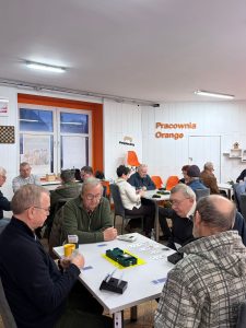 Na zdjęciu znajduję się grupa osób grająca w brydża przy stole.