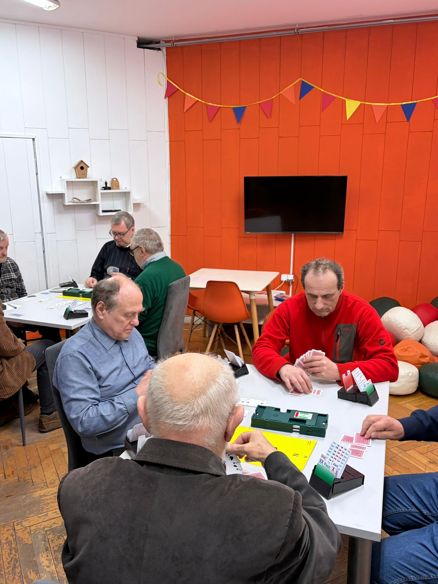 Na zdjęciu znajduję się grupa osób grająca w brydża przy stole.