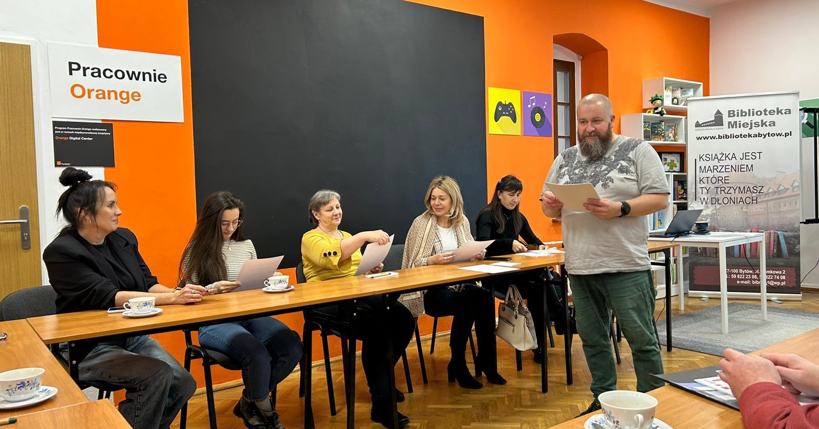 Grupa osób siedzi przy stolikach, przed nimi stoi prowadzący.