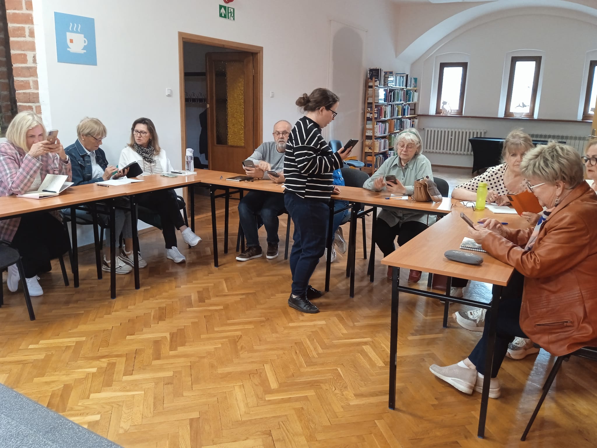 Grupa osób siedzi przy stolikach podczas lekcji języka angielskiego.