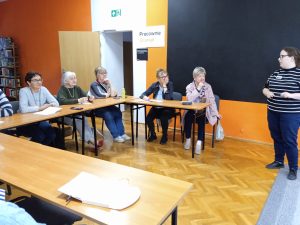 Grupa kobiet siedzi przy stolikach, przed nimi stoi prowadząca zajęcia.