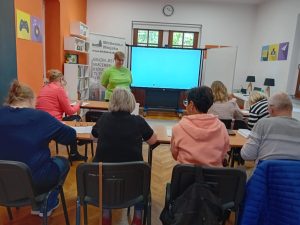 Grupa osób siedzących na krzesłach. Przed nimi prezentacja wyświetlona na ekranie.