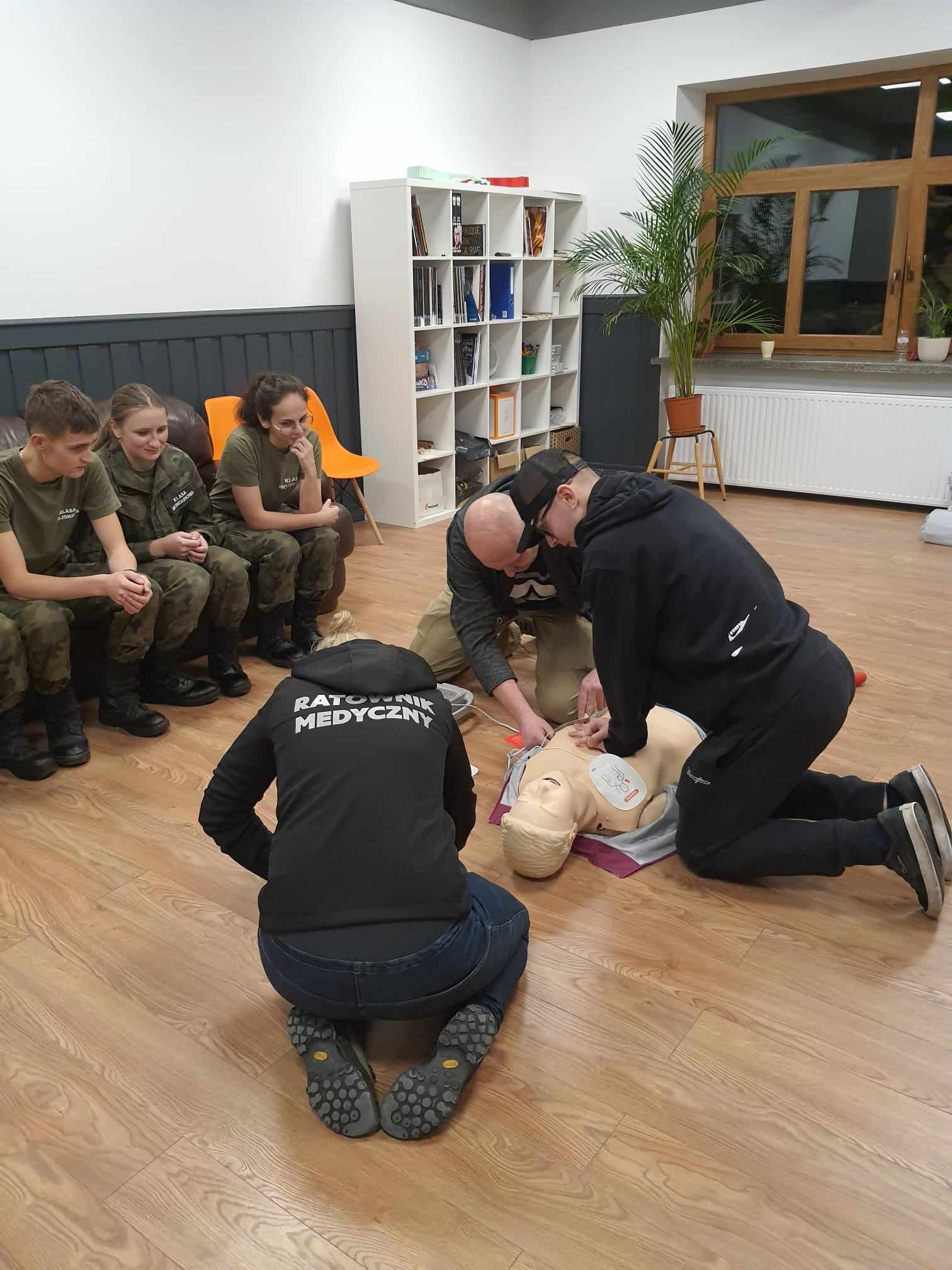 Dwóch mężczyzn na przemian uciska klatkę piersiową manekina, słuchając wskazówek ratownika.