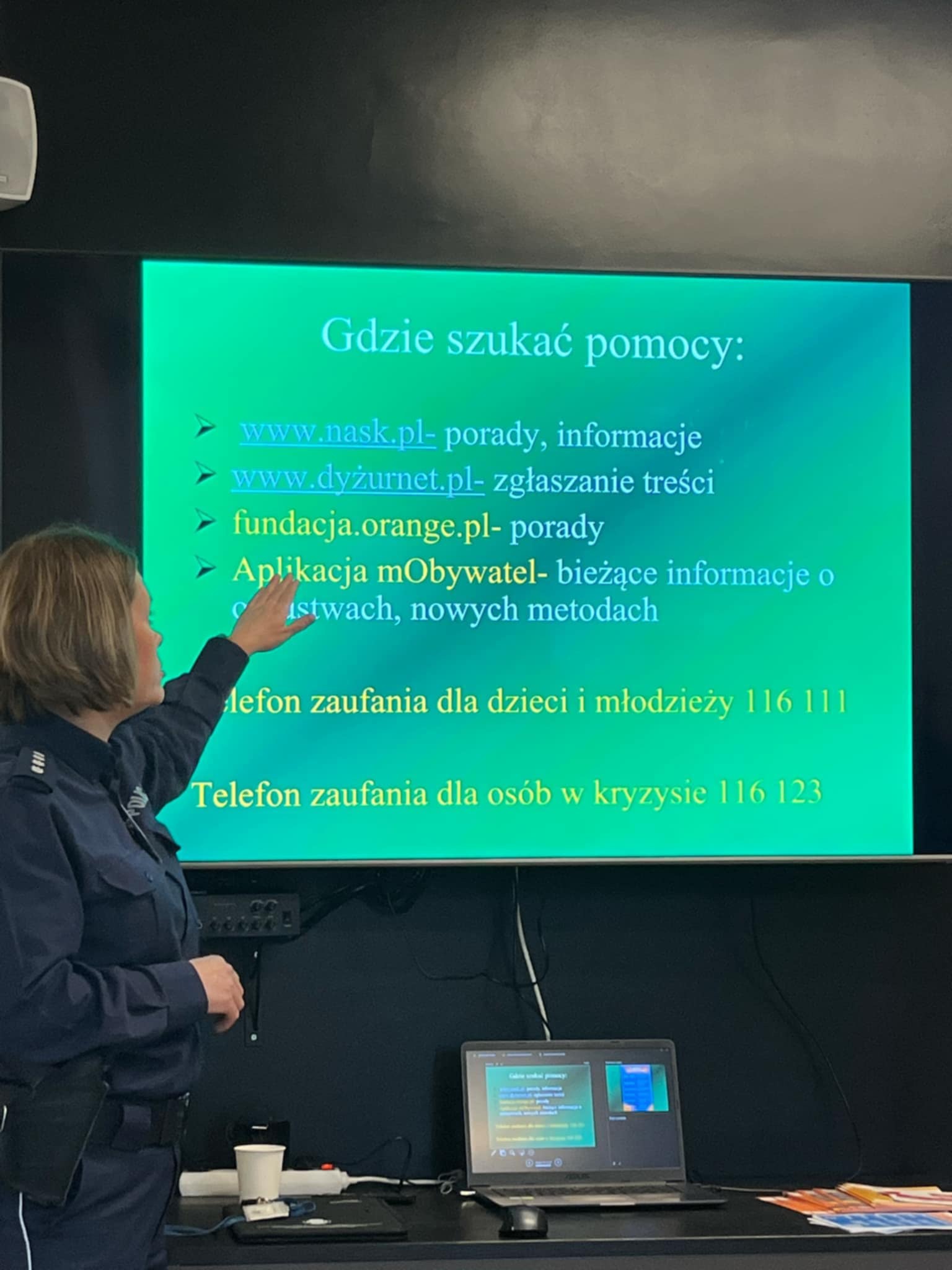 Policjantka wskazuje na ekranie telewizora organizacje gdzie można szukać pomocy przy zagrożeniach wynikających z internetu