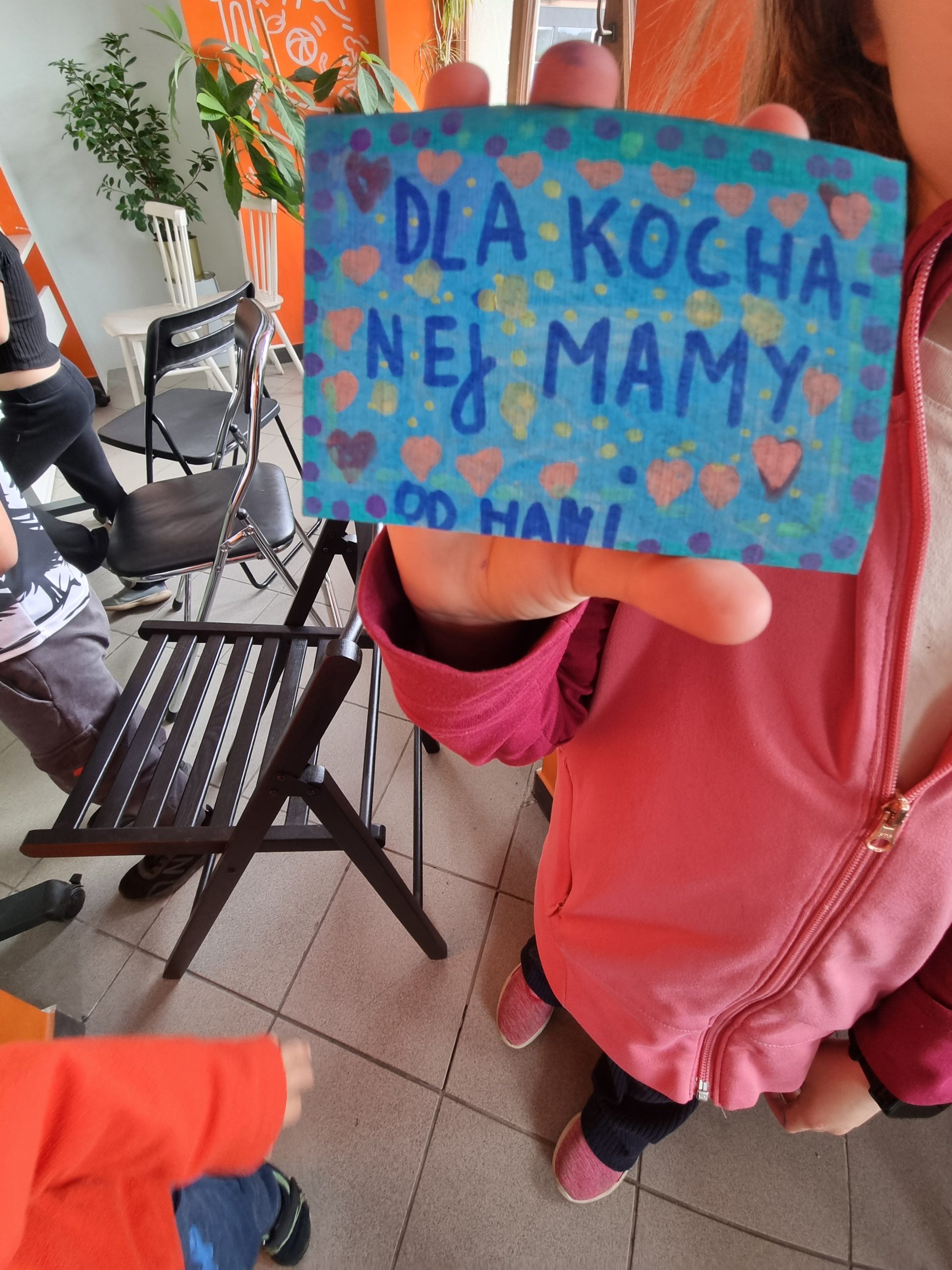 Dziewczynka trzyma w ręce, pomalowany na niebiesko magnes na lodówkę. Jest na nim napis - dla kochanej mamy.